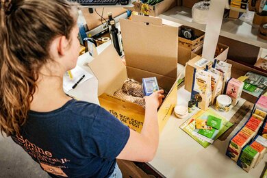 The photo shows an employee carefully packing the ordered products into a SONNENTOR shipping carton by hand. | © SONNENTOR