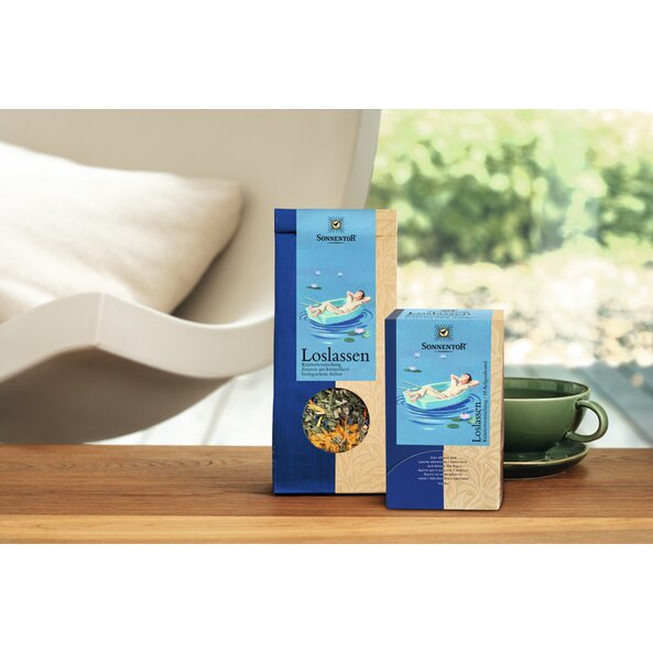 Photo of a pack of Let it Be Herbal Tea on a table, left of it is a pack of Let it Be Herbal Tea loose and right a green cup.