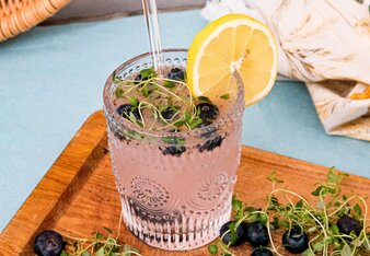 Ein schönes Glas steht auf einem Brett, gefüllt mit dem SONNENTOR Tschin Tschin Sirup, Heidelbeeren und frischem Thymian, garniert mit einer Zitronenscheibe.  | © SONNENTOR