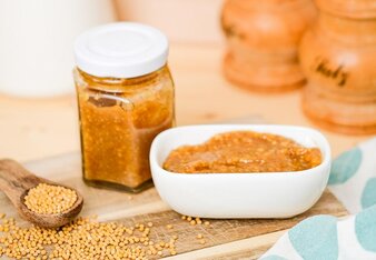 Der Feigen-Senf ist ein einem Glas abgefüllt und befindet sich ebenso in einem weißen länglichen Schüsserl daneben. Außerdem ist ein Holzlöffel mit Senfkörnern zu sehen, welche auch dazwischen lose verteilt sind.  | © SONNENTOR