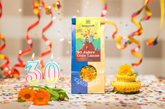 30 Jahre Gute Laune | © SONNENTOR Auf dem Foto sieht man eine Packung 30 Jahre Gute Laune Tee. Daneben sieht man Kerzen Geburtstagszahlen, Konfetti und Luftschlangen. | © SONNENTOR