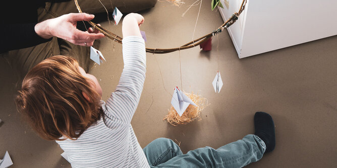 Papier Hühner Mobile | © SONNENTOR Auf dem Foto sieht man wie ein Kind die selbst gebastelten Hühner auf einen Ring hängt. | © SONNENTOR