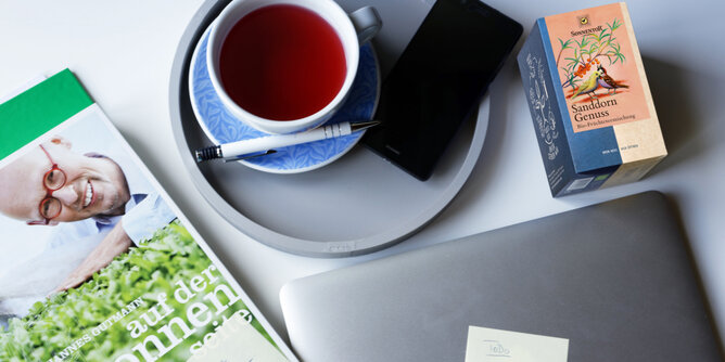 Tee around the clock | © SONNENTOR Auf dem Foto sieht man eine Tasse Sanddorn Tee. Auf dem Foto ist auch ein Buch, ein Stift, ein Handy und ein Laptop zu sehen. | © SONNENTOR