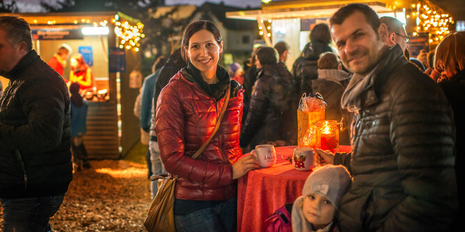Adventmarkt | © SONNENTOR Auf dem Foto sieht man eine Familie auf einem Adventmarkt. Die Eltern halten jeweils eine SONNENTOR Tasse in den Händen. | © SONNENTOR