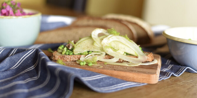 Belegtes Brot mit Apfel, Erbsen und Fenchel | © SONNENTOR Auf dem Foto sieht man ein Holzbrett mit einer Scheibe Brot darauf. Das Brot ist mit Apfel, Erbsen und Fenchel belegt. | © SONNENTOR