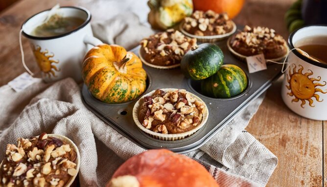 Nudelholz Kürbis | © SONNENTOR Auf dem Foto sind Kürbismuffins zu sehen. Sie sind mit Nüssen garniert. Daneben sieht man kleine Kürbisse und zwei Tassen Tee. | © SONNENTOR