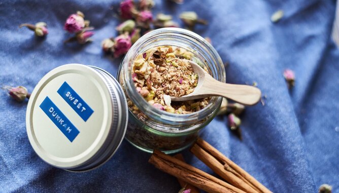 Süße Dukkah | © SONNENTOR Auf dem Foto sieht man ein Glas mit süßem Dukkah. Daneben liegen Zimtstangen und Rosenblütenknospen. | © SONNENTOR