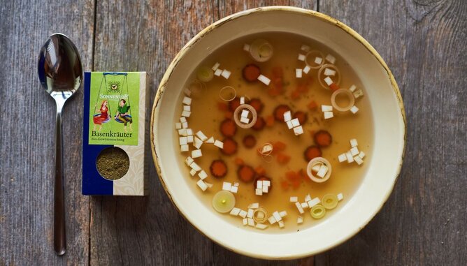 Nudelholz Basensuppe | © SONNENTOR Auf dem Foto sieht man eine klare Basensuppe mit Suppengemüse darin. Neben dem Suppenteller liegt eine Packung Basenkräuter. | © SONNENTOR