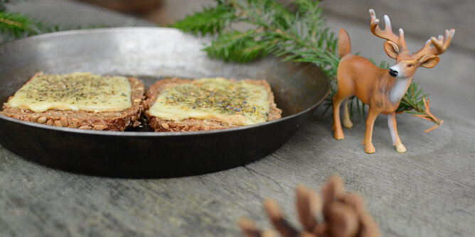 Raclette-Brot | © SONNENTOR Auf dem Foto sind zwei Raclette Brote zu sehen. Daneben sieht man Zweige, Zapfen und einen Schleich Hirsch. | © SONNENTOR